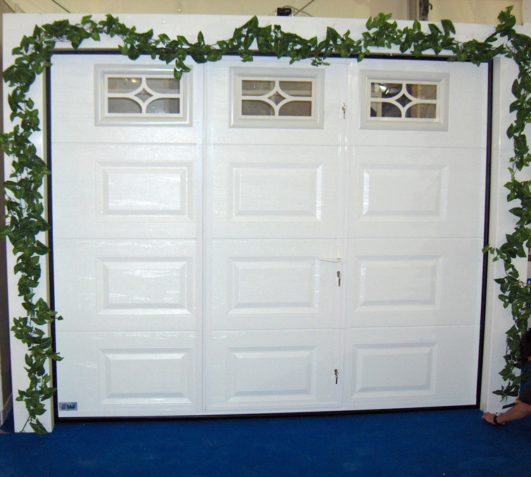 Habitat cassette portillon entouré de végétation avec porte de garage sectionnelle blanche en pvc et trois fenêtres ajourées pour les particuliers.